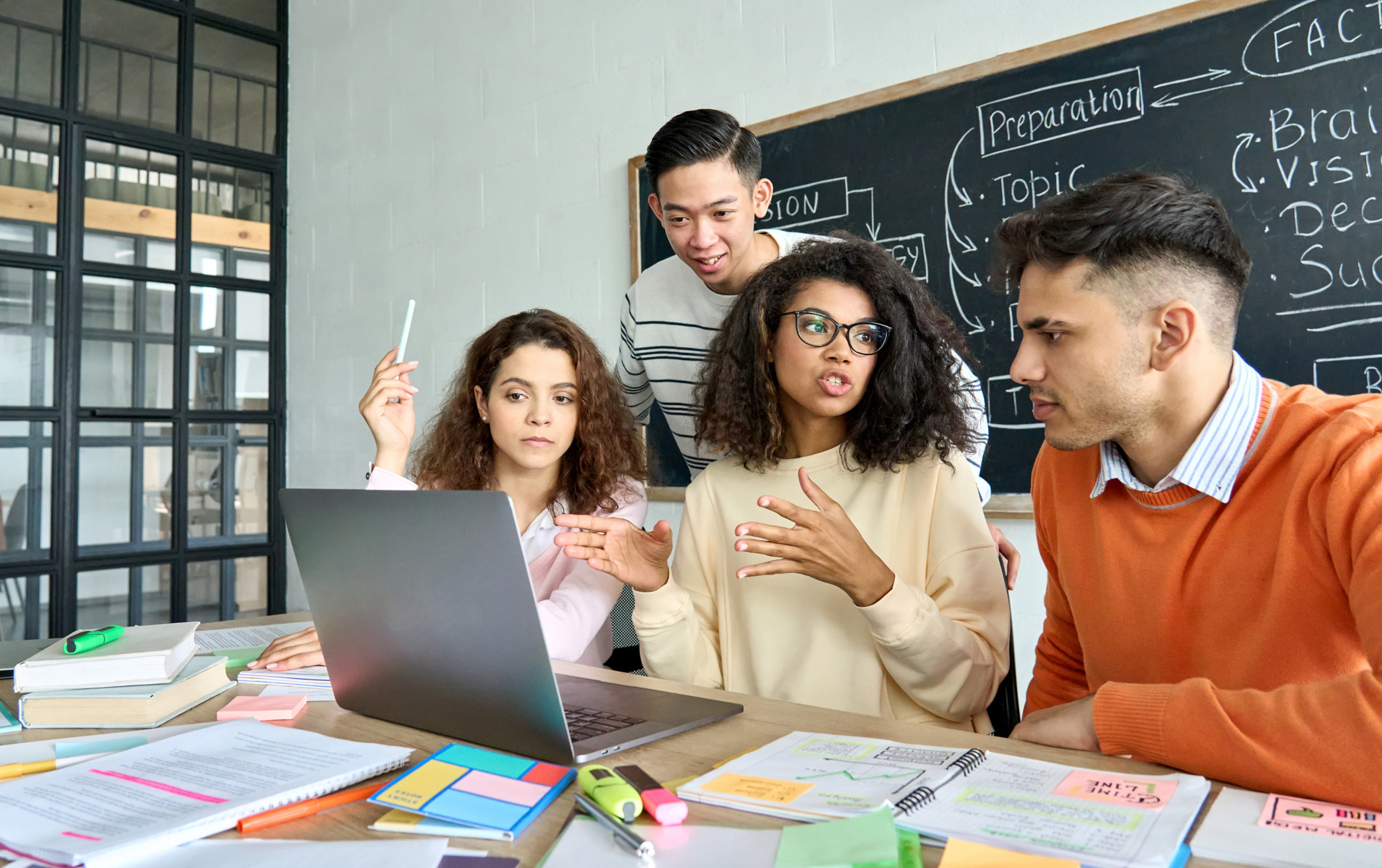 iCivics curridylum: Four older students sit around laptop at table covered in multi-colored note papers in active discussion