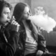 youth tobacco use, vaping: Black and white photo of two older teens sitting against brick wall smoking electronic cigarettes