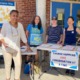 Mass. graduation requirements: Group of four ethnically diverse middle age women stand outside in a covered walkway at a table with info signs